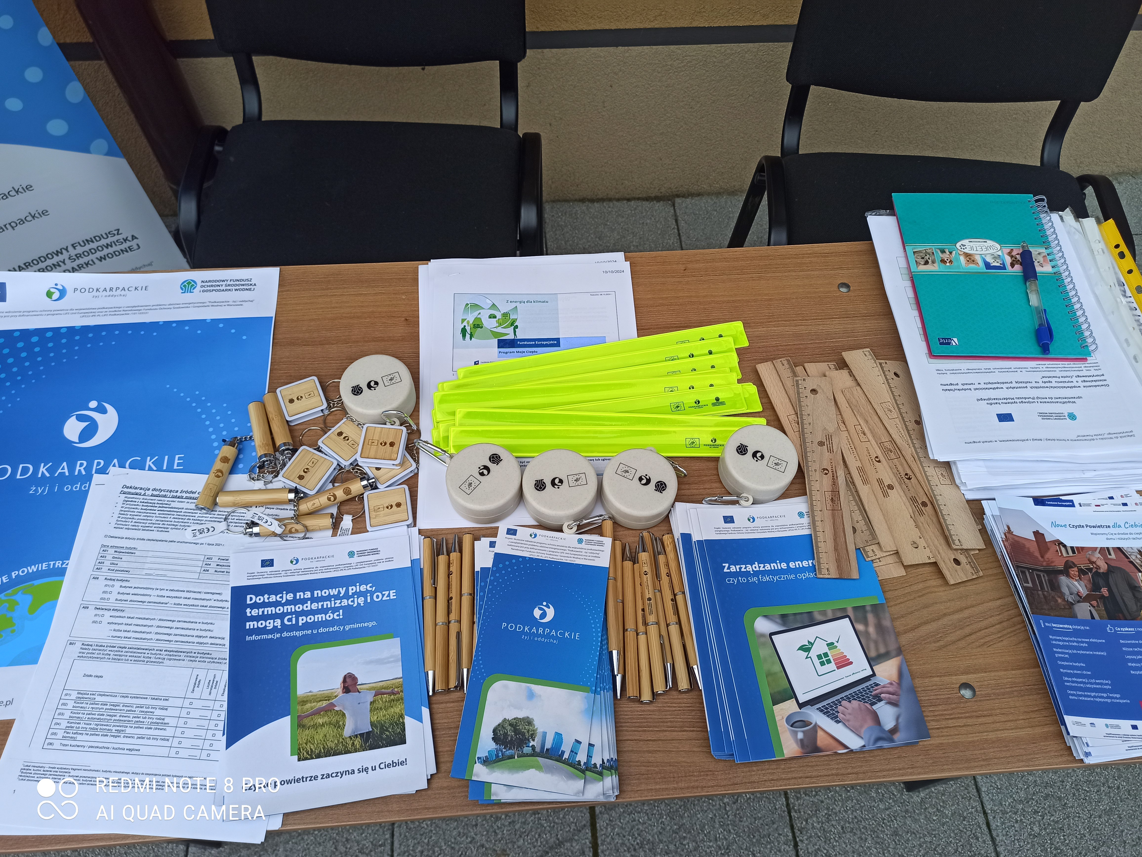 St&oacute;ł z materiałami promocyjnymi: ulotki, długopisy, miarki, odblaski, informujące o programach ekologicznych Podkarpacia.